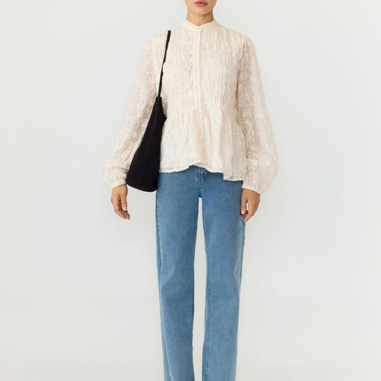 A model stands against a plain light gray studio background wearing a cream lace long-sleeve blouse, light blue straight-leg jeans, black pointed shoes, and carrying a black shoulder bag.