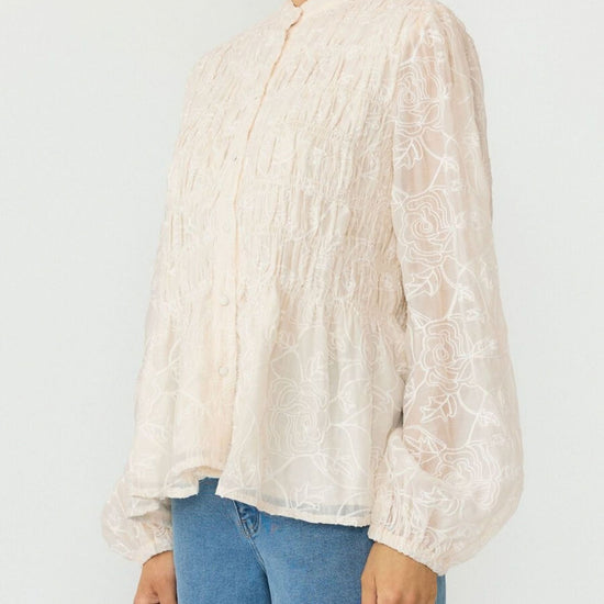 A woman wearing a cream-colored, long-sleeved lace blouse with a gathered smocked bodice and blue jeans, shown from the chin down against a plain light background.