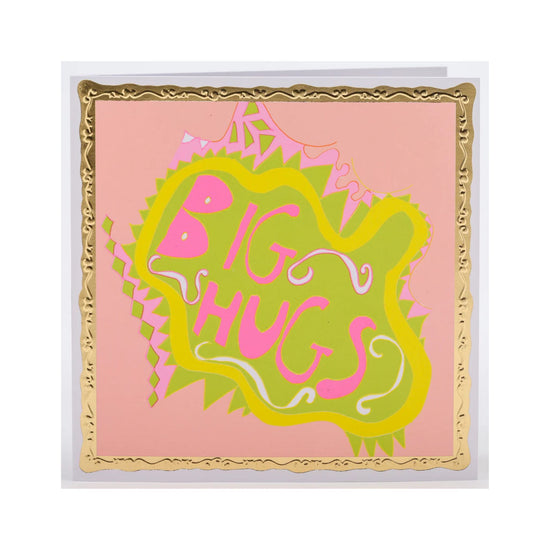 A square greeting card featuring a pink background with a decorative gold border and a central green and pink abstract shape that reads 'BIG HUGS'.
