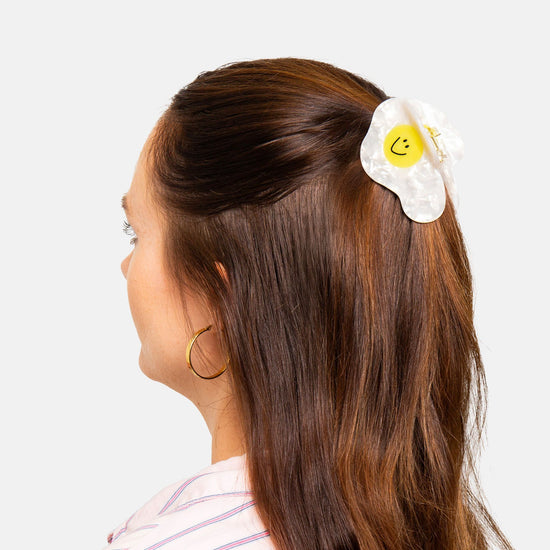 Person with long brown hair wearing a white fried egg shaped hair clip with a yellow smiley face as yolk on a light gray background