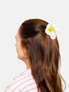 Person with long brown hair wearing a white fried egg shaped hair clip with a yellow smiley face as yolk on a light gray background