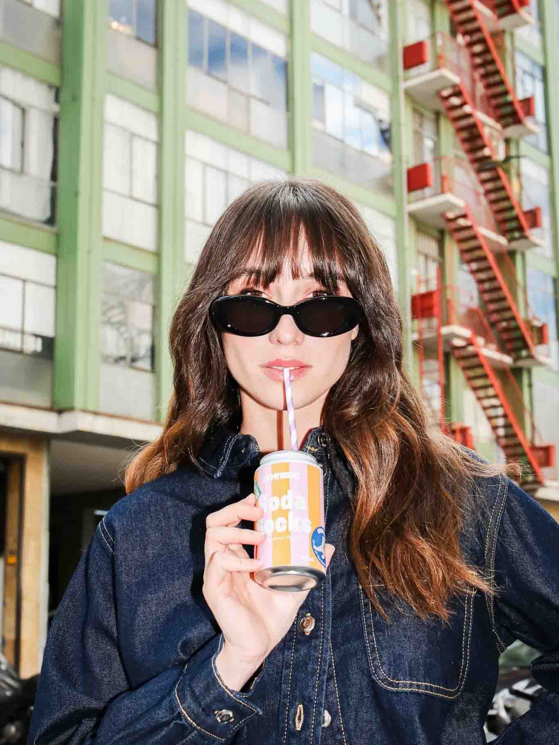 Woman pretending to drink from a soda can with a straw in an urban setting