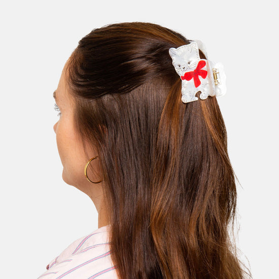 Person with long brown hair wearing a white cat shaped hair clip with a red bow on a light gray background