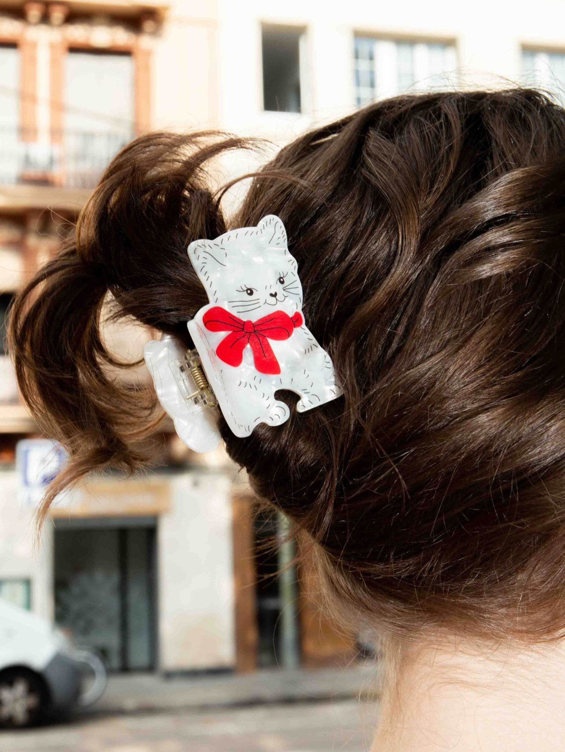 Hair clip with a cat design and red bow in a person's hair, blurred city street background