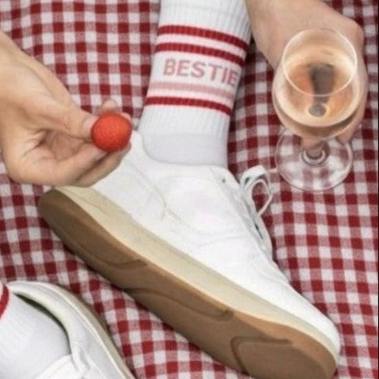 White sneakers with brown soles on a red and white checkered fabric, person holding a glass of pink drink and a small red object.