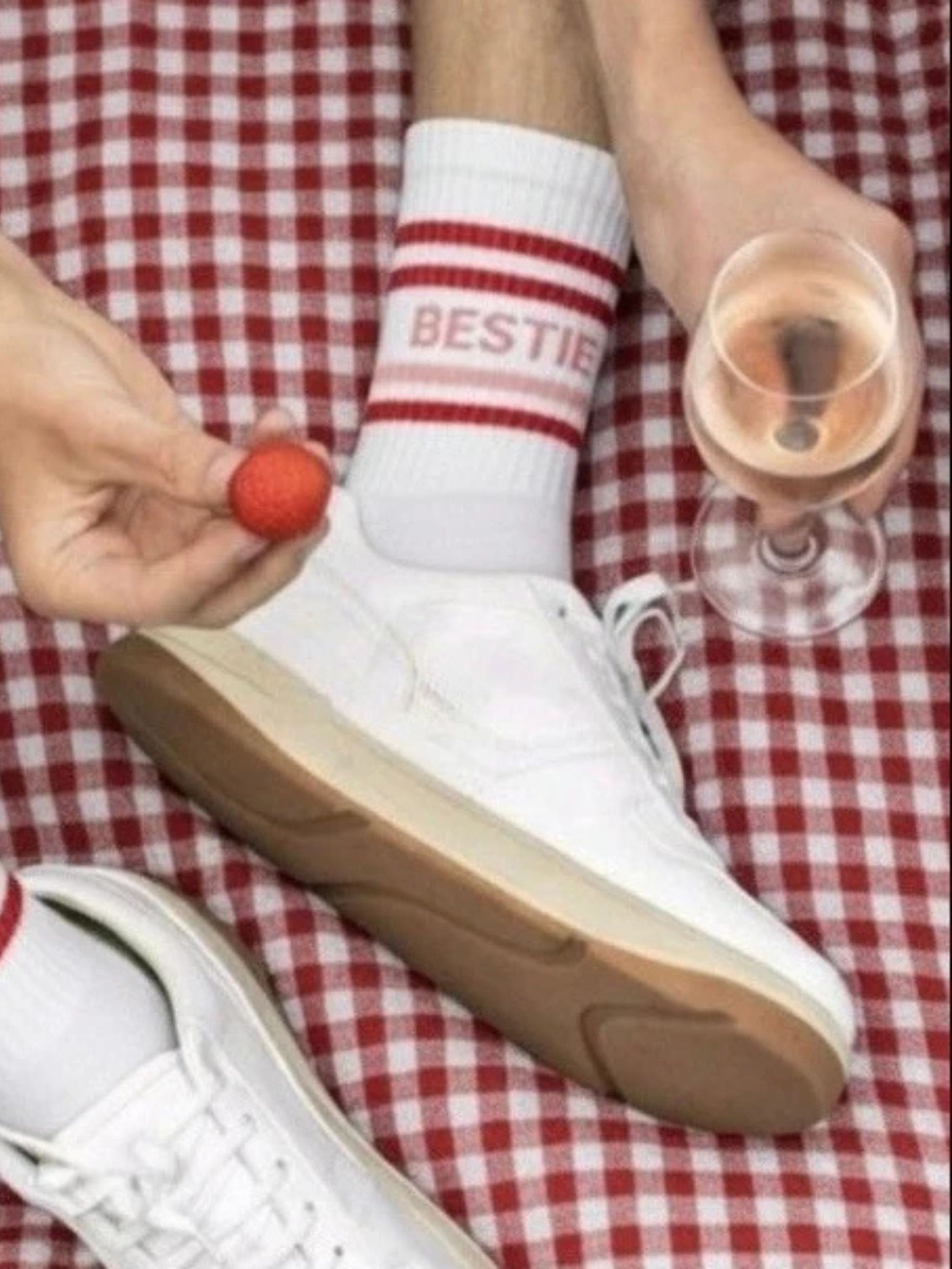 White sneakers with brown soles on a red and white checkered fabric, person holding a glass of pink drink and a small red object.