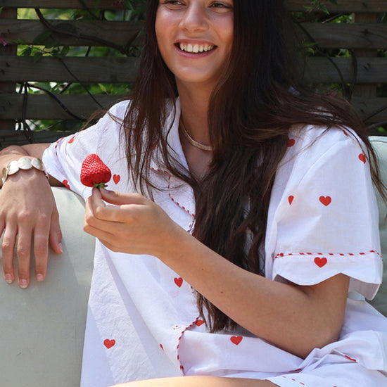 A woman sitting outdoors wearing a white short-sleeve shirt and shorts set with small red heart prints and red dashed trim, holding a strawberry.
