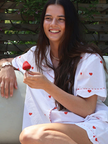 A woman sitting outdoors wearing a white short-sleeve shirt and shorts set with small red heart prints and red dashed trim, holding a strawberry.