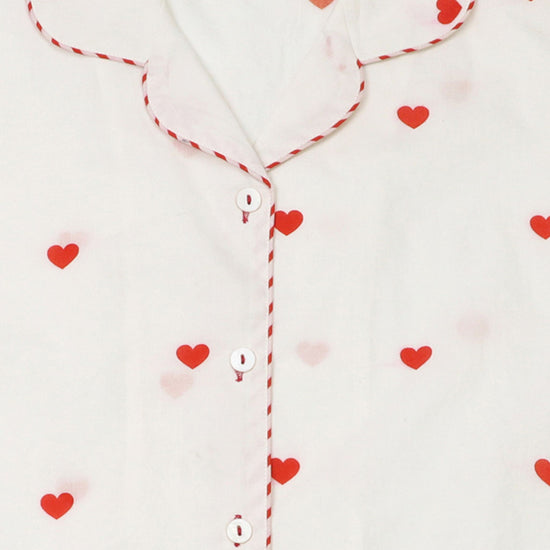 Close-up of a white pyjama top with red heart prints, red-and-white striped piping along the collar and placket, and white buttons.