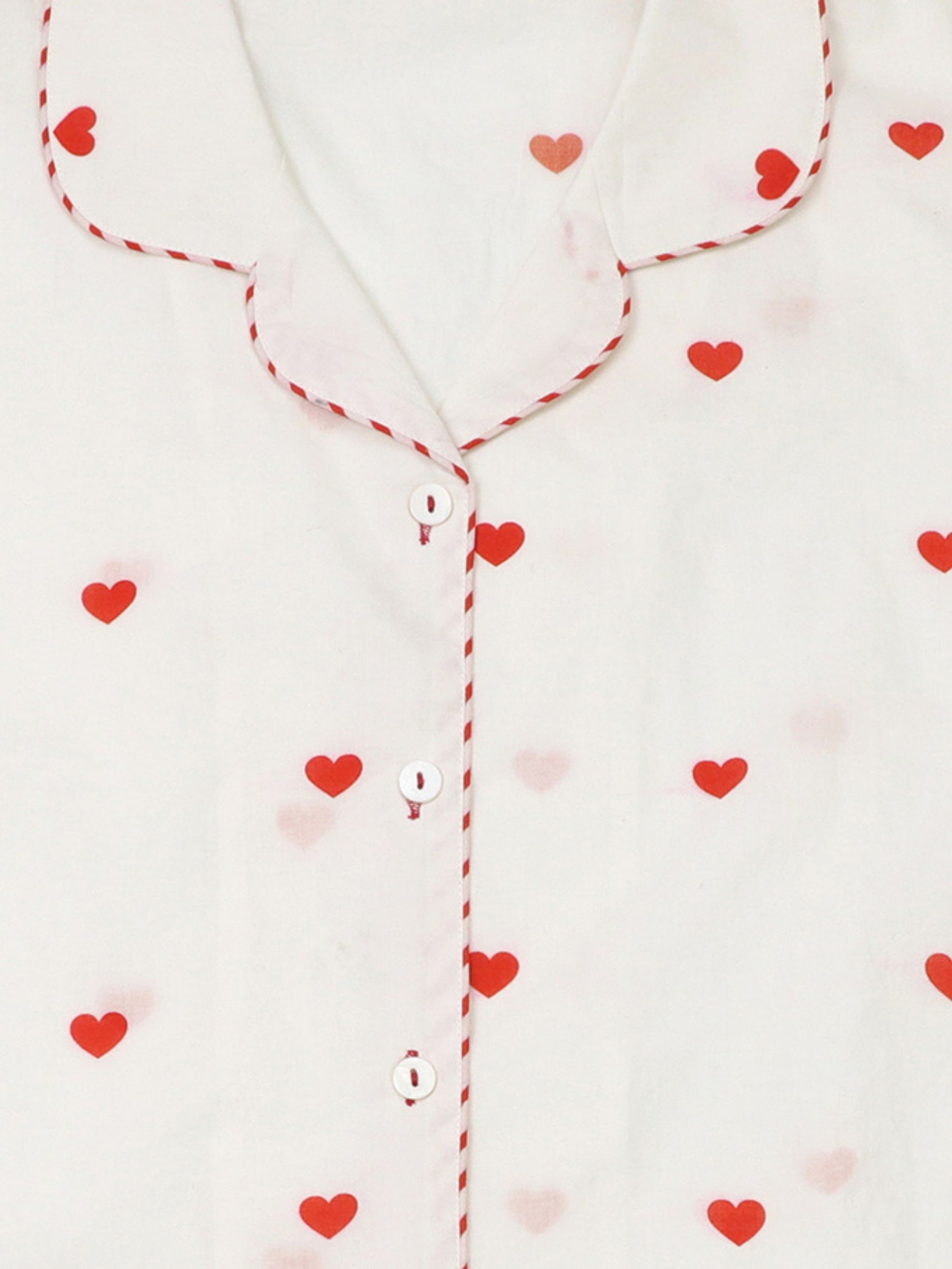 Close-up of a white pyjama top with red heart prints, red-and-white striped piping along the collar and placket, and white buttons.