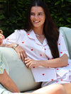 A woman smiling while wearing a white short-sleeve pajama set with red heart prints and red piped trim, seated on a light-colored outdoor lounge chair with green foliage in the background.